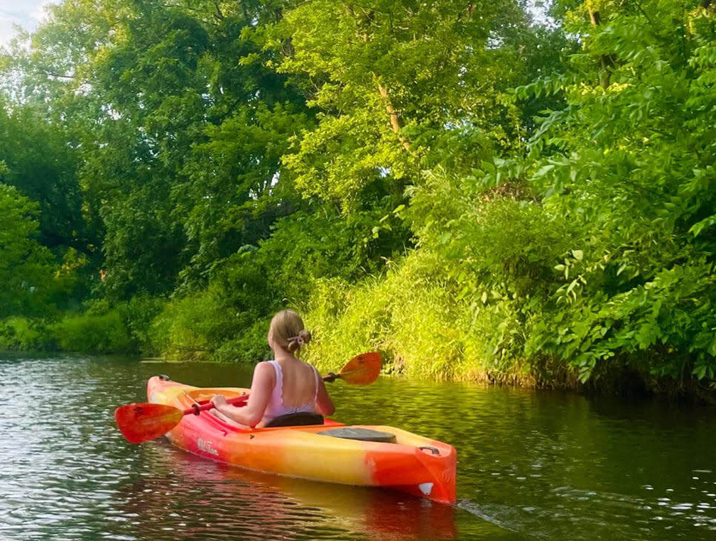 A woman on the water in a kayak. She's surrounded by a very bright and vivid forest scene.