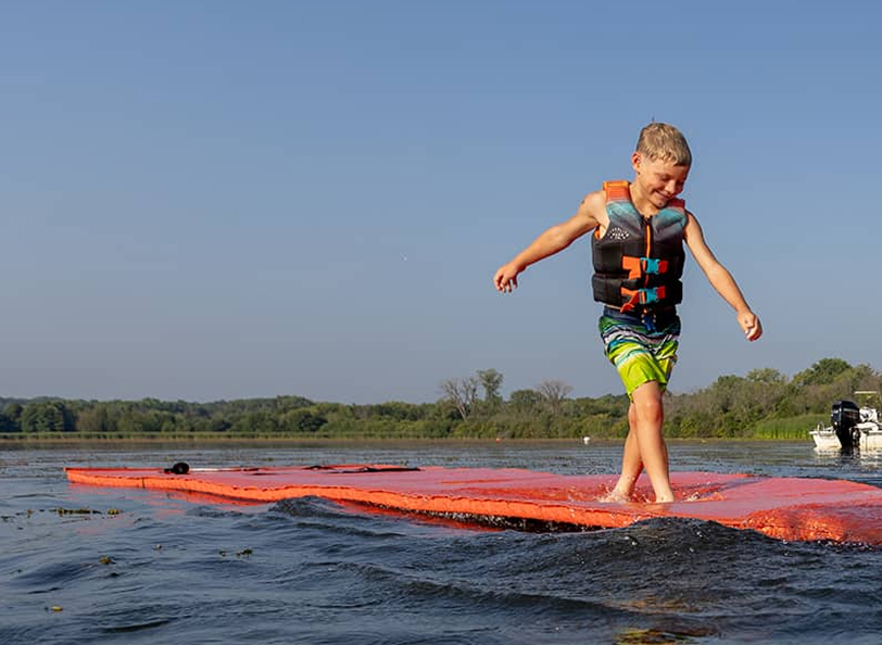 Kid in a life jacket standing on a watermat.